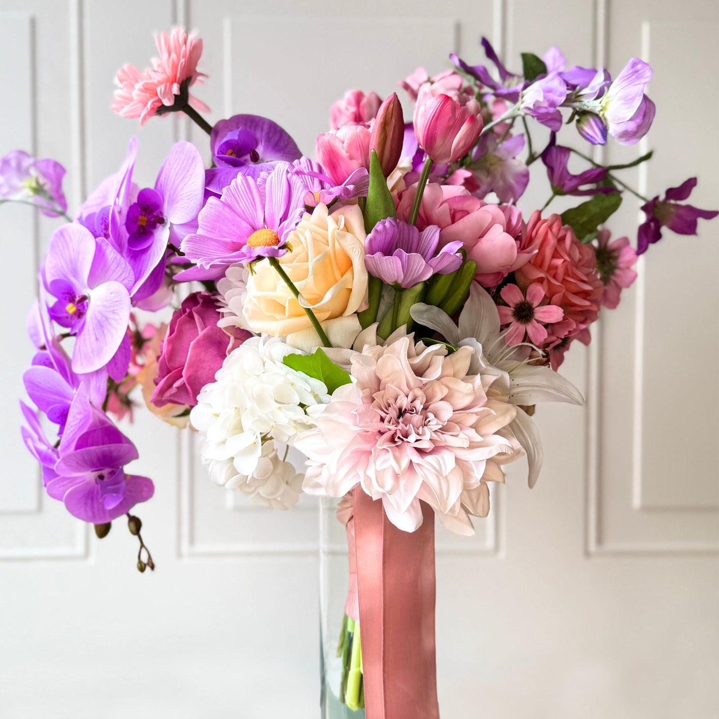 artificial flower bridal bouquet featuring purple artificial orchids, pink artificial cosmos, champagne artificial tea roses, mauve artificial dahlia, coral artificial gerbera, light pink artificial Austin roses, coral artificial tea roses, deep pink artificial peonies, and white artificial hydrangeas, tied with a peach ribbon — from My Faux Florist Canada.