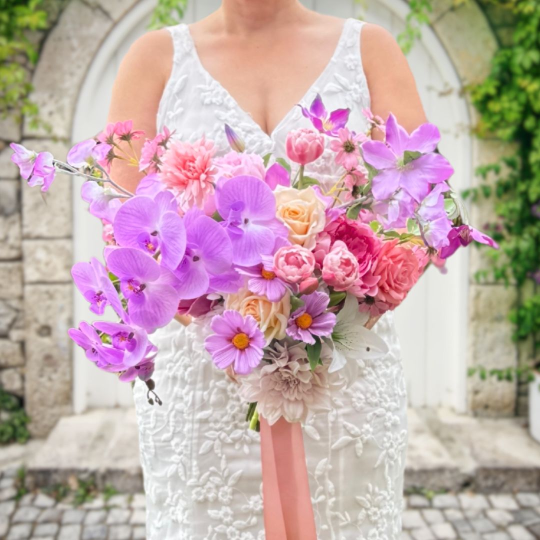 Bride holding a realistic artificial flower bridal bouquet featuring purple artificial orchids, pink artificial cosmos, champagne artificial tea roses, mauve artificial dahlia, coral artificial gerbera, light pink artificial Austin roses, coral artificial tea roses, deep pink artificial peonies, and white artificial hydrangeas, tied with a peach ribbon — from My Faux Florist Canada.