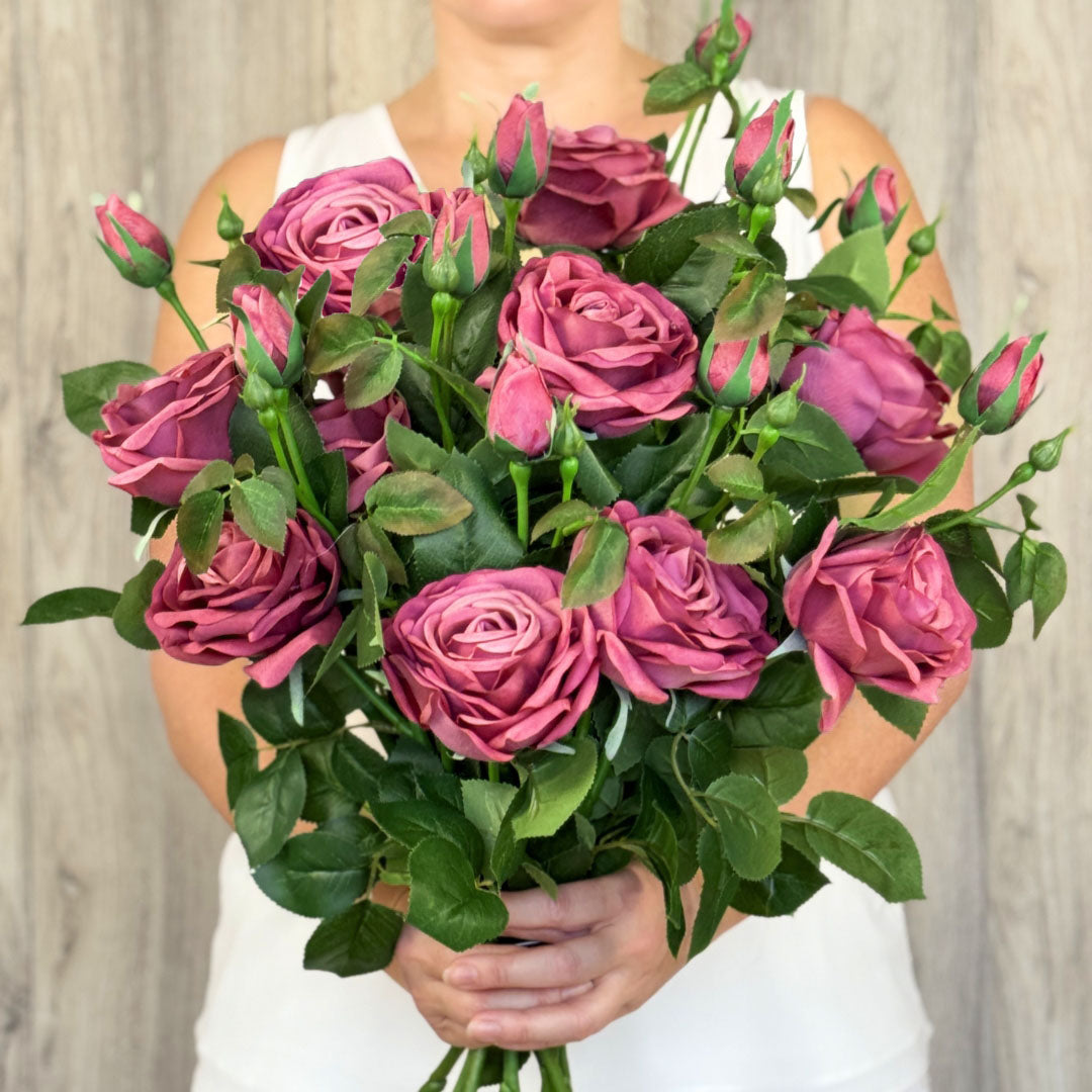 A person holding an burgundy realistic artificial rose spray with fully open and closed buds, and green leaves by My Faux Florist Canada
