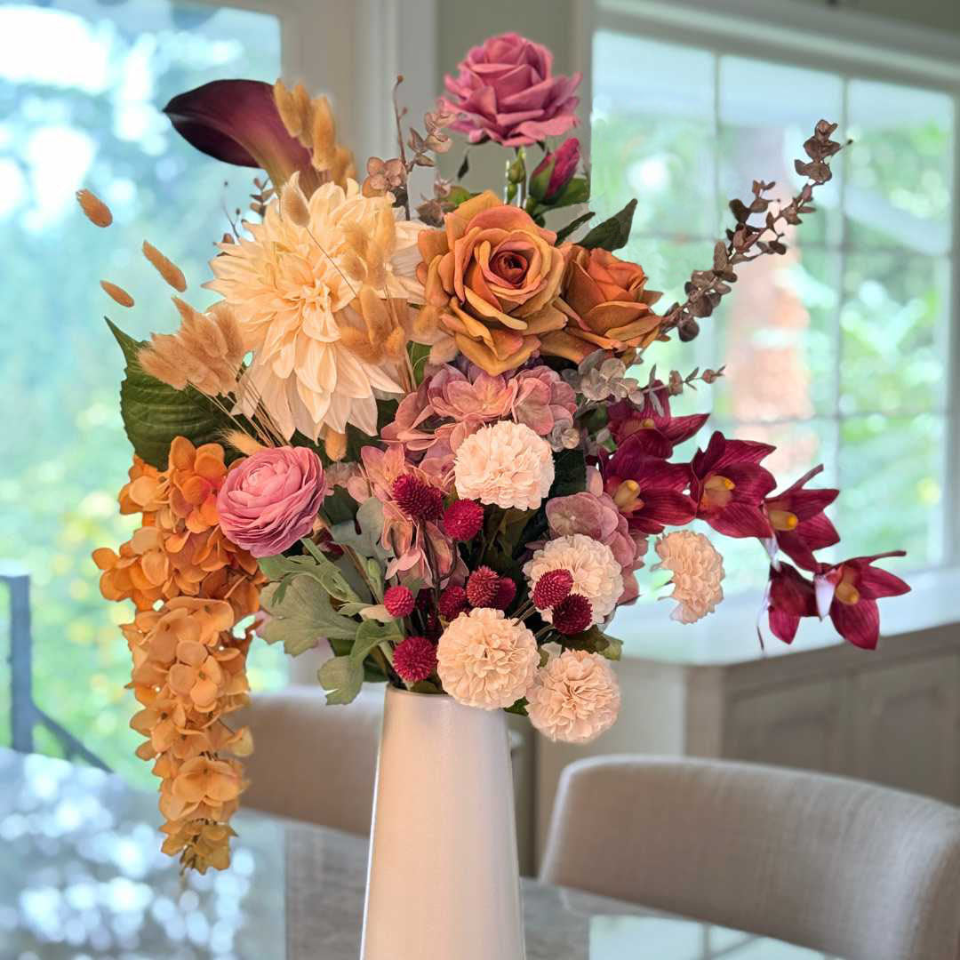 Realistic artificial flower arrangement in a tall white vase featuring cream artificial dahlias, apricot artificial roses, mauve artificial roses, pink artificial ranunculus, blush artificial hydrangeas, burgundy artificial orchids, red artificial billy balls, and golden artificial hanging orchids — from My Faux Florist Canada.