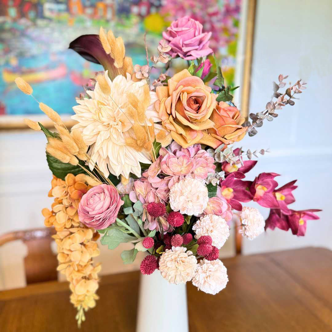 Realistic artificial flower arrangement in a tall white vase featuring cream artificial dahlias, apricot artificial roses, mauve artificial roses, pink artificial ranunculus, blush artificial hydrangeas, burgundy artificial orchids, red artificial billy balls, and golden artificial hanging orchids — from My Faux Florist Canada.
