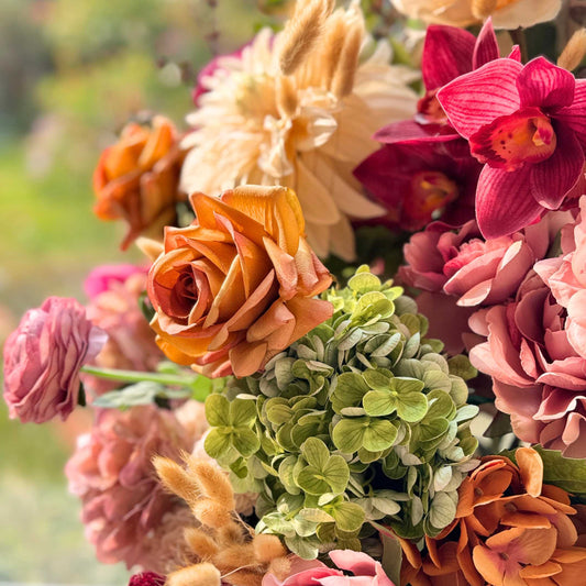 Realistic artificial flower arrangement in a white vase featuring cream artificial dahlias, apricot artificial roses, mauve artificial roses, pink artificial hydrangeas, blush artificial peonies, green artificial hydrangeas, burgundy artificial ranunculus, and golden artificial orchids — from My Faux Florist Canada.