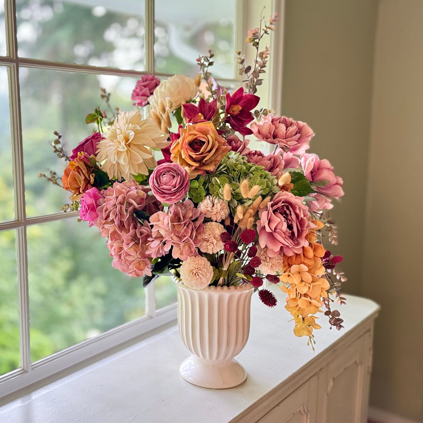 Realistic artificial flower arrangement in a white vase featuring cream artificial dahlias, apricot artificial roses, mauve artificial roses, pink artificial hydrangeas, blush artificial peonies, green artificial hydrangeas, burgundy artificial ranunculus, and golden artificial orchids — from My Faux Florist Canada.