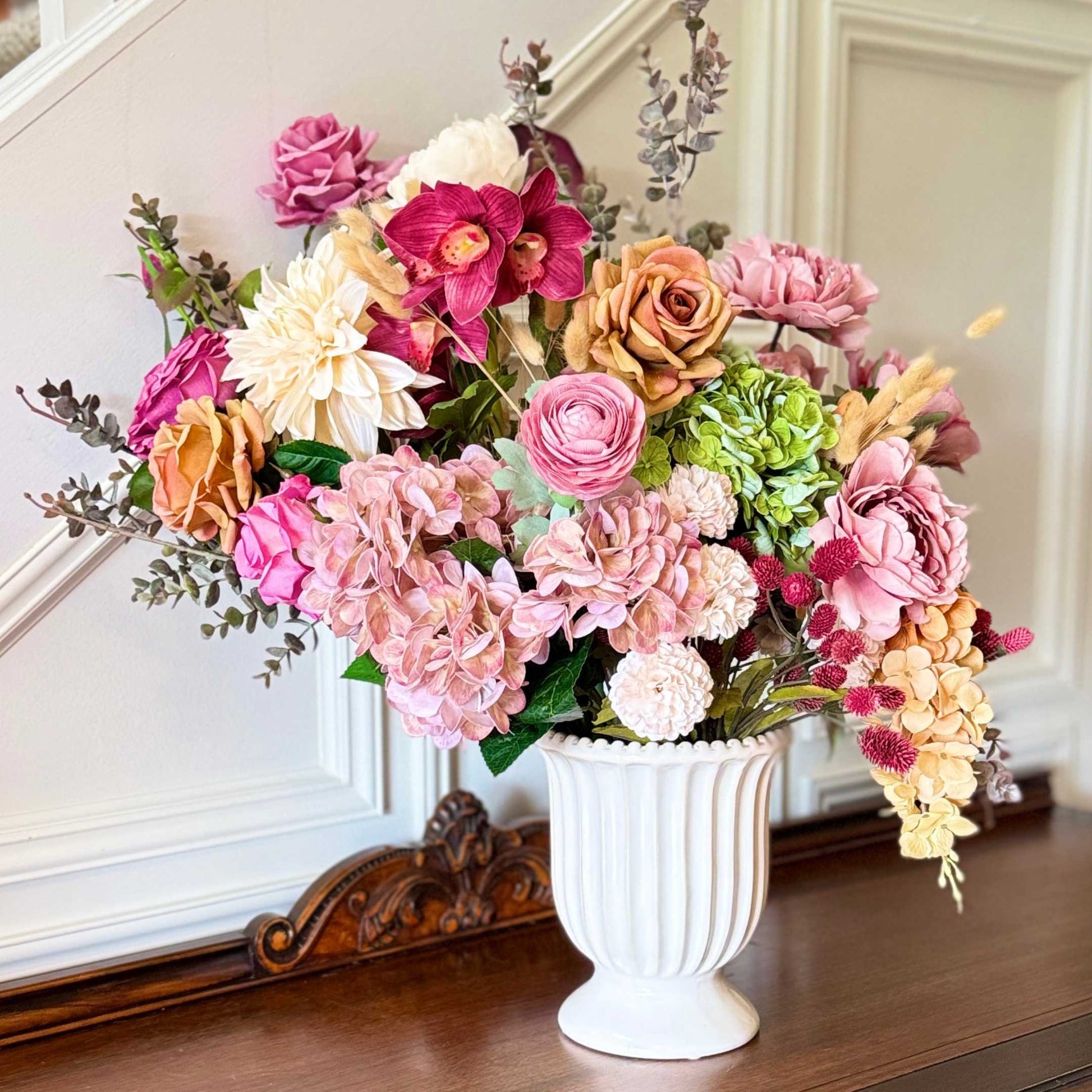 Realistic artificial flower arrangement in a white vase featuring cream artificial dahlias, apricot artificial roses, mauve artificial roses, pink artificial hydrangeas, blush artificial peonies, green artificial hydrangeas, burgundy artificial ranunculus, and golden artificial orchids — from My Faux Florist Canada.