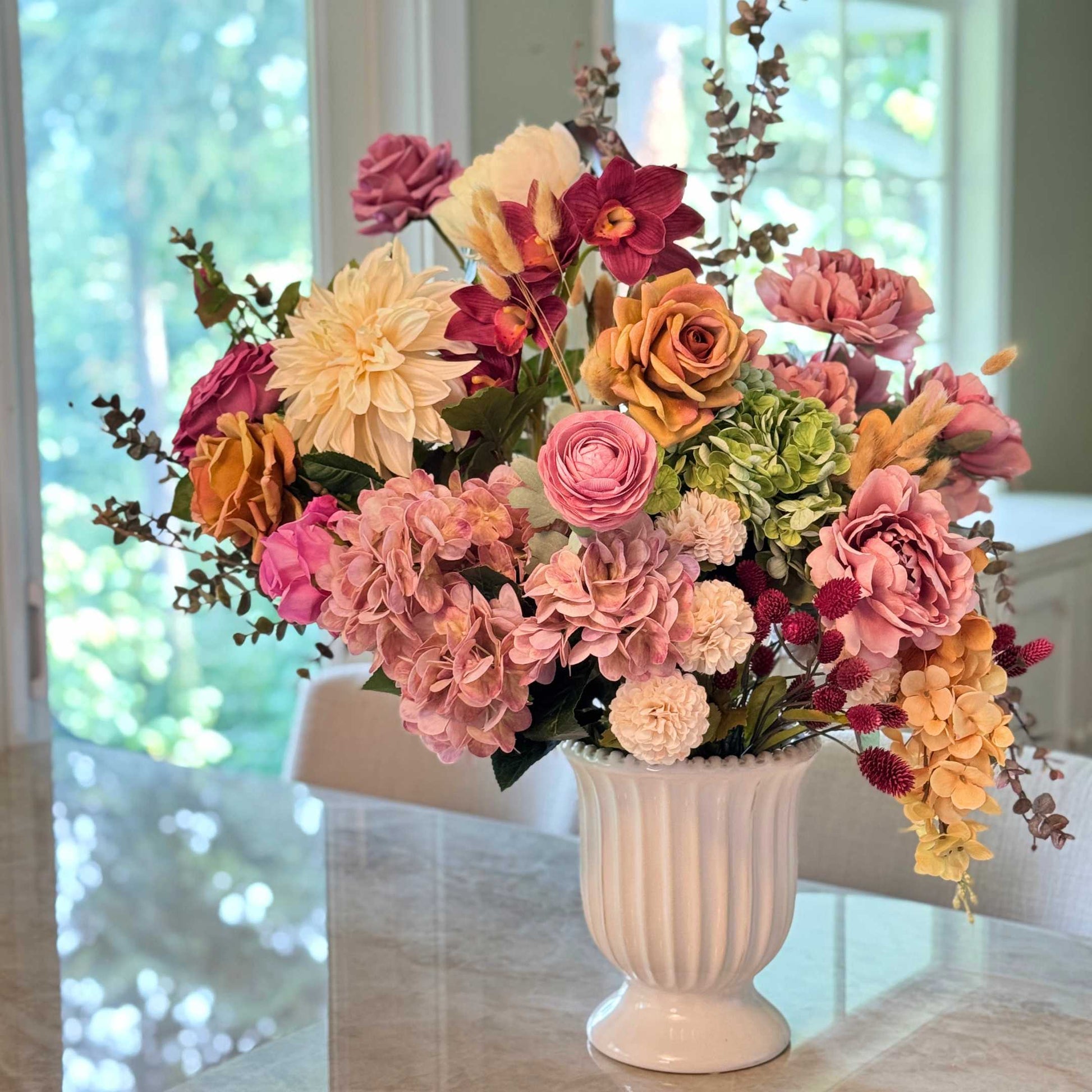 Realistic artificial flower arrangement in a white vase featuring cream artificial dahlias, apricot artificial roses, mauve artificial roses, pink artificial hydrangeas, blush artificial peonies, green artificial hydrangeas, burgundy artificial ranunculus, and golden artificial orchids — from My Faux Florist Canada.