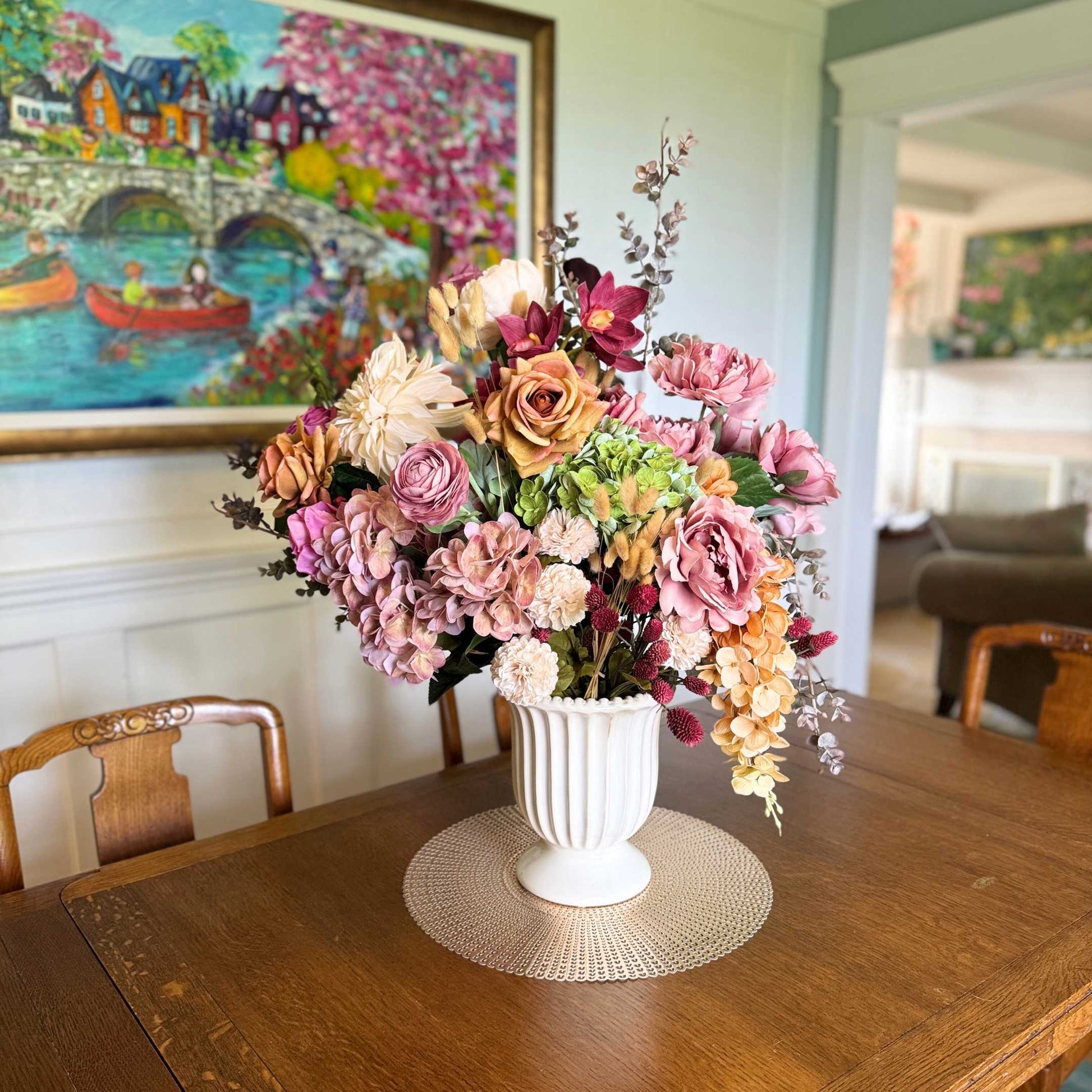 Realistic artificial flower arrangement in a white vase on a table featuring cream artificial dahlias, apricot artificial roses, mauve artificial roses, pink artificial hydrangeas, blush artificial peonies, green artificial hydrangeas, burgundy artificial ranunculus, and golden artificial orchids — from My Faux Florist Canada.