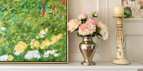 Alt text: Realistic artificial flower arrangement in a silver vase featuring blush artificial roses, ivory artificial roses, soft pink artificial peonies, and white artificial hydrangeas, styled on a mantel beside a green floral painting and cream candlestick — from My Faux Florist Canada.