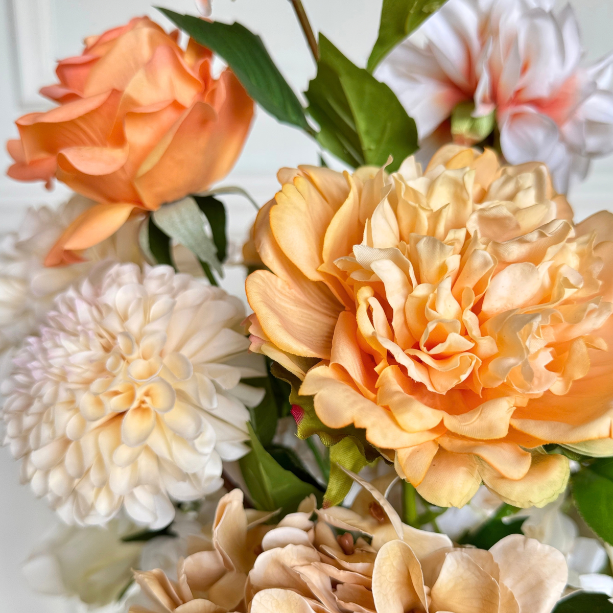 A close-up of realistic artificial flowers, featuring a large peach Real Touch peony in the center, a white faux dahlia on the left, and other soft peach and cream blooms with green leaves. For My Faux Florist Canada.