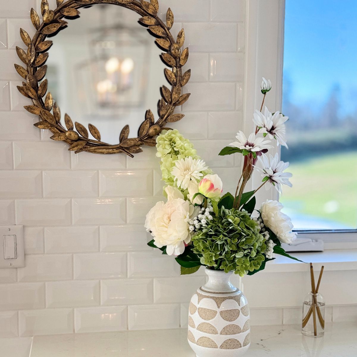 Decorative mirror with floral arrangement of realistic artificial flowers on a table against a tiled wall. For My Faux Florist Canada.