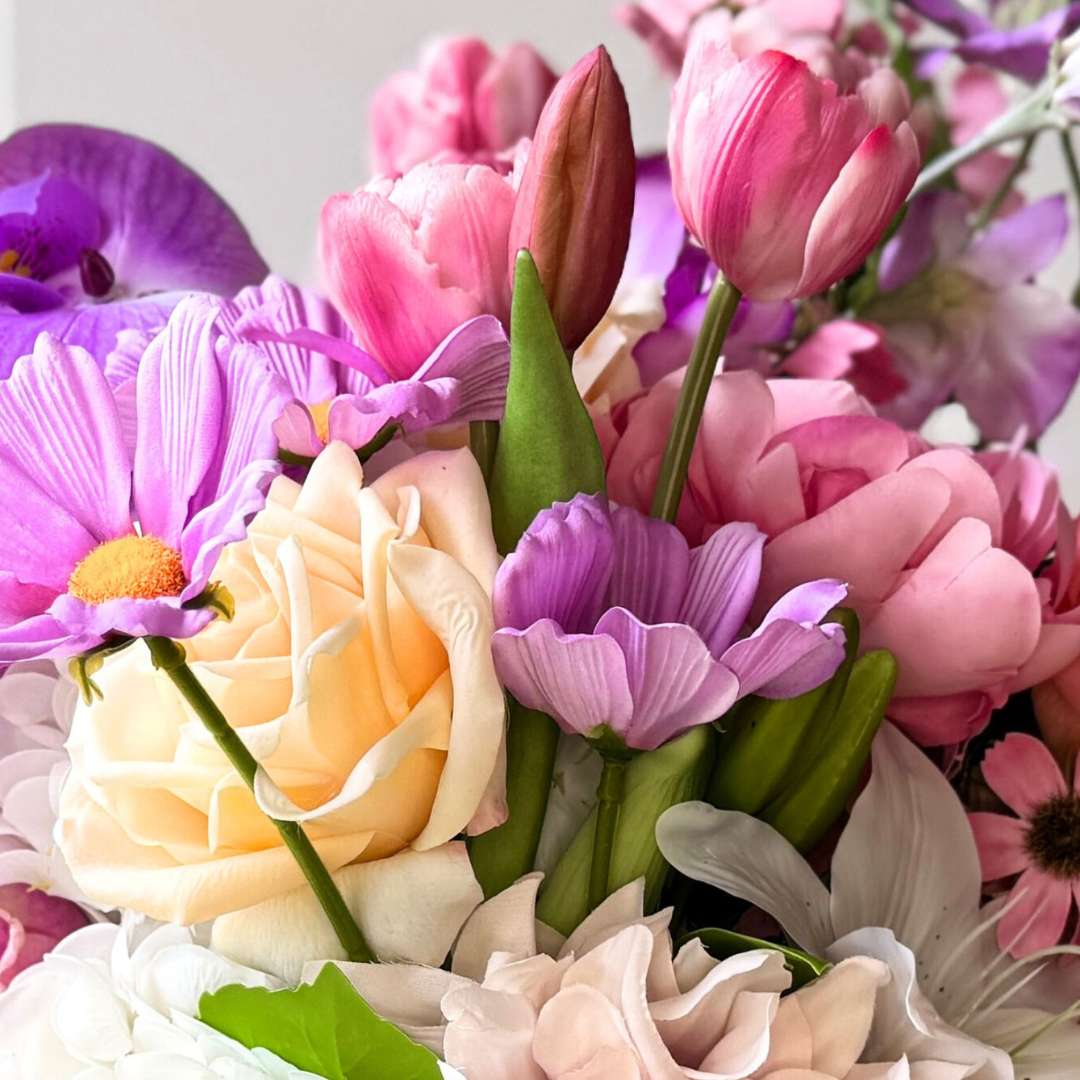 close up of an artificial flower bridal bouquet featuring purple artificial orchids, pink artificial cosmos, champagne artificial tea roses, mauve artificial dahlia, coral artificial gerbera, light pink artificial Austin roses, coral artificial tea roses, deep pink artificial peonies, and white artificial hydrangeas, tied with a peach ribbon — from My Faux Florist Canada.