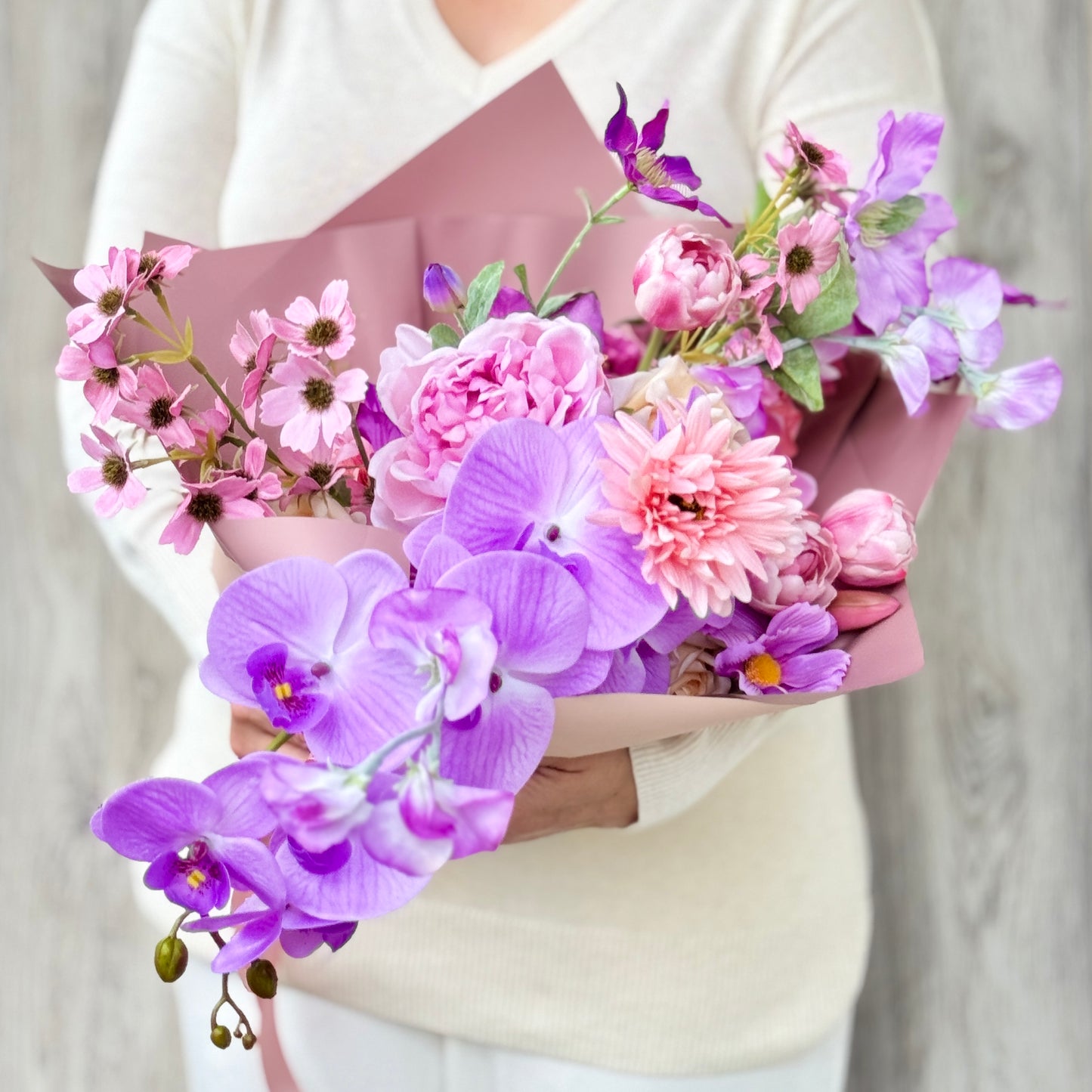 Bouquet of pink and purple realistic artificial flowers held by a person wearing a white sweater. Faux peonies, faux roses, faux hydrangea, and faux orchids in an artificial flower bouquet for My Faux Florist Canada.