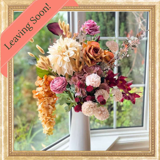 Realistic artificial flower arrangement in a tall white vase featuring cream artificial dahlias, apricot artificial roses, mauve artificial roses, pink artificial ranunculus, blush artificial hydrangeas, burgundy artificial orchids, red artificial billy balls, and golden artificial hanging orchids — from My Faux Florist Canada.