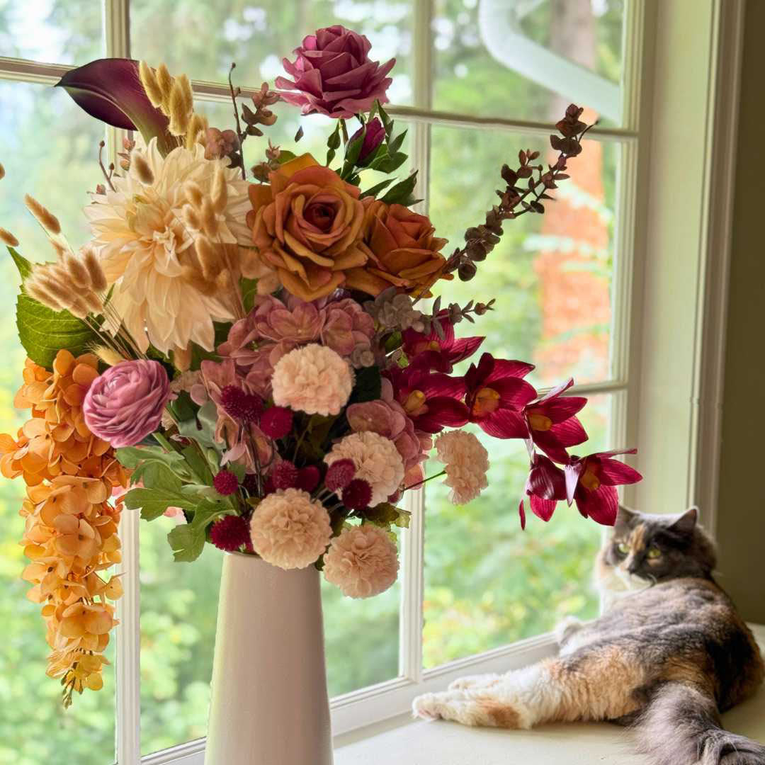 Realistic artificial flower arrangement in a tall white vase featuring cream artificial dahlias, apricot artificial roses, mauve artificial roses, pink artificial ranunculus, blush artificial hydrangeas, burgundy artificial orchids, red artificial billy balls, and golden artificial hanging orchids — from My Faux Florist Canada.