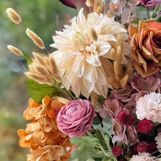 Realistic artificial flower arrangement in a tall white vase featuring cream artificial dahlias, apricot artificial roses, mauve artificial roses, pink artificial ranunculus, blush artificial hydrangeas, burgundy artificial orchids, red artificial billy balls, and golden artificial hanging orchids — from My Faux Florist Canada.