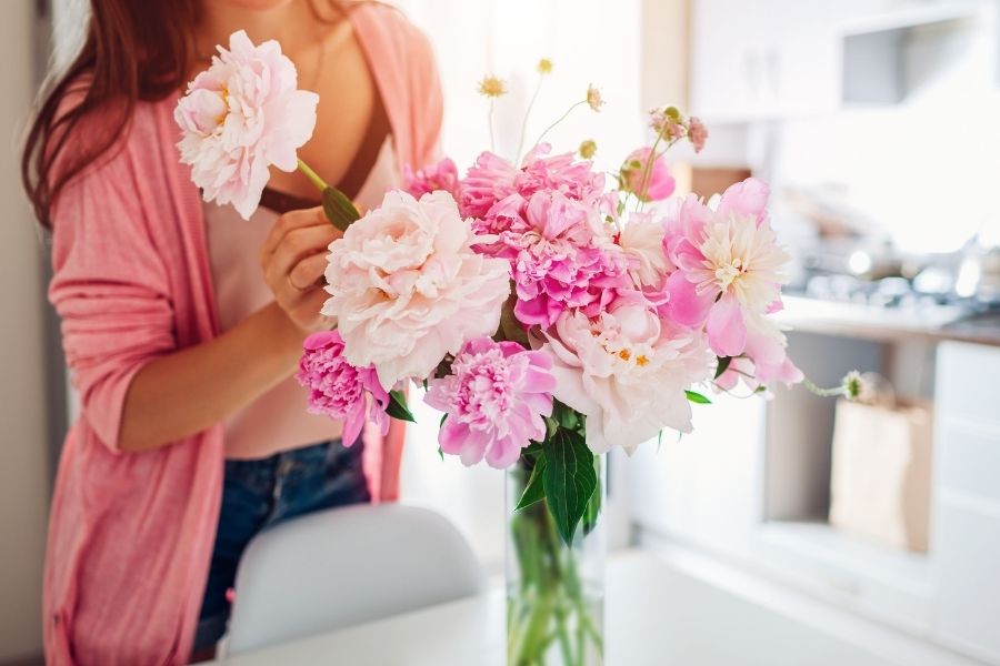 woman arranging a bouquet of fake flowers in shades of pink in a clear vase on a kitchen counter