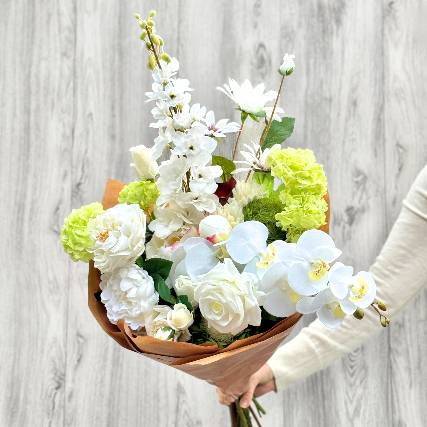 Bouquet of white and green realistic artificial flowers held by a person against a light gray wooden background. Faux peonies, faux roses, faux hydrangea, and faux orchids in an artificial flower bouquet for My Faux Florist Canada.