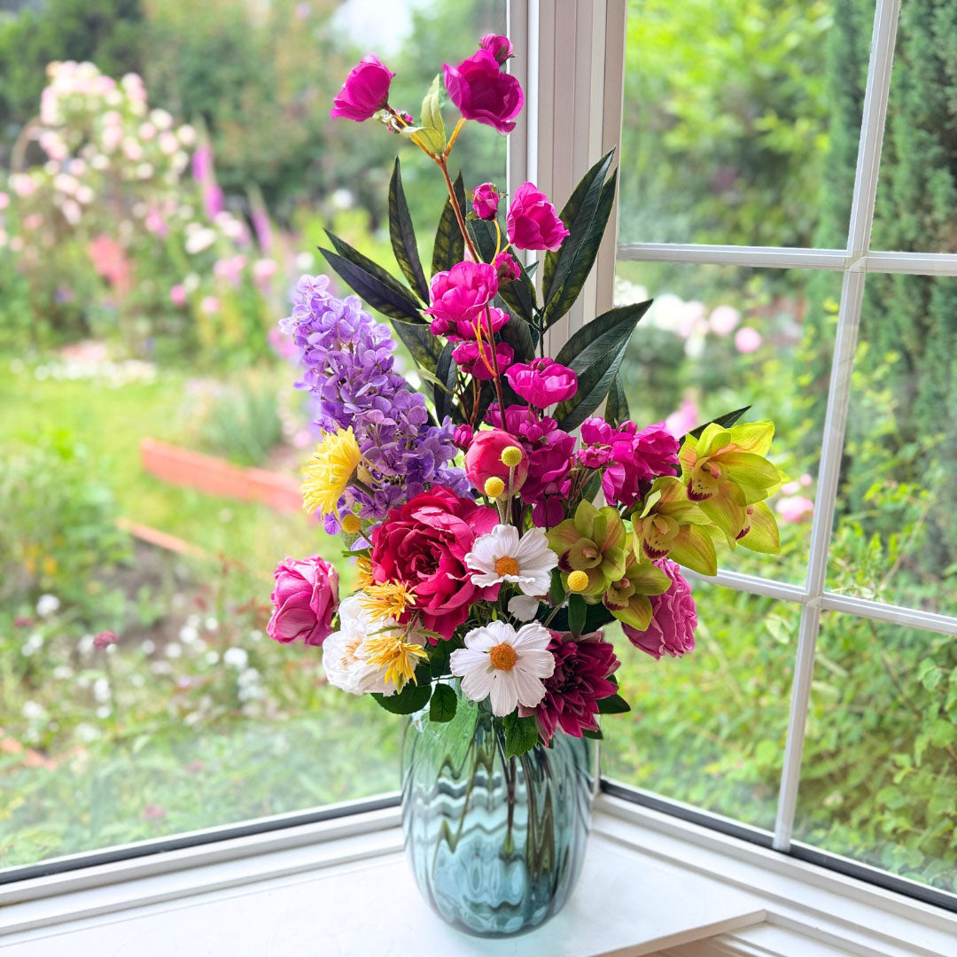 Realistic artificial flower arrangement in a blue glass vase featuring fuchsia artificial orchids, purple artificial delphiniums, green artificial cymbidium orchids, white artificial cosmos, yellow artificial dahlias, pink artificial roses, and magenta artificial peonies — from My Faux Florist Canada.