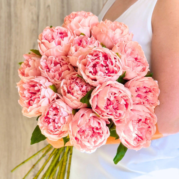 Artificial flower bouquet featuring pink faux peonies held by a woman in a white dress.