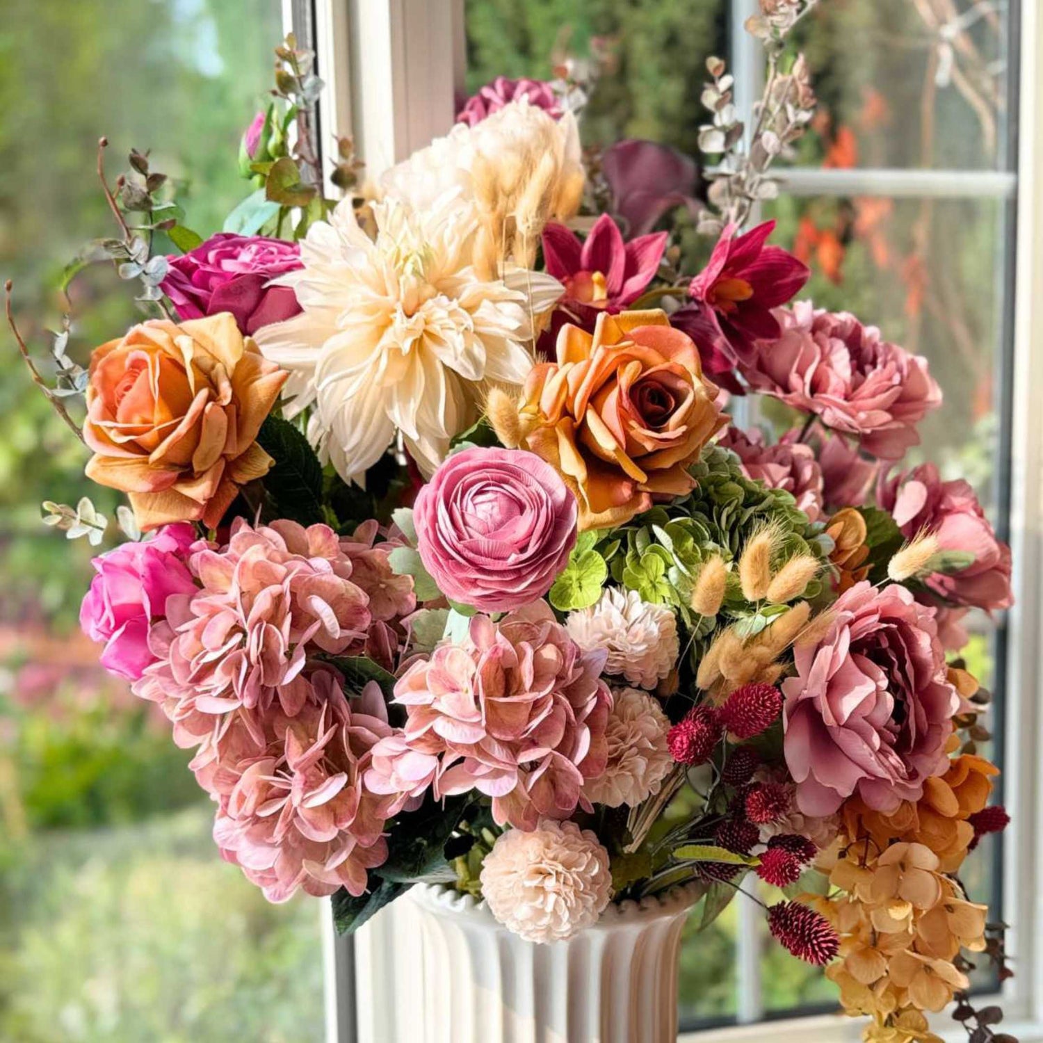 Vase full of lifelike artificial flowers in a window in a variety of muted fall colours from fake roses and artificial dahlias to artificial orchids and artificial wisteria