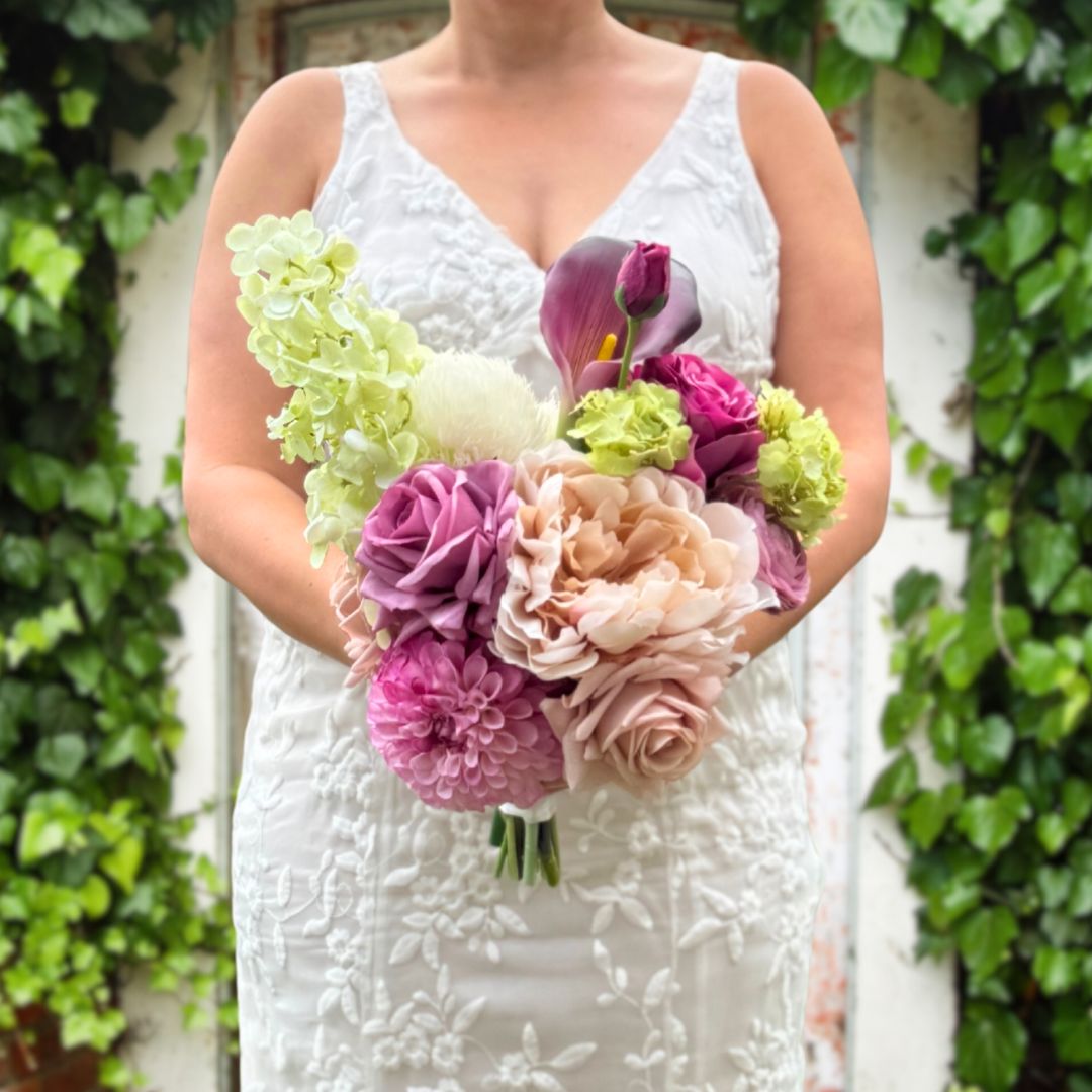 Bride holding a realistic artificial flower bridal bouquet featuring antique cream artificial roses, dusty mauve artificial roses, blush artificial peonies, pink artificial dahlias, pale green artificial hydrangeas, green artificial peonies, and deep wine artificial calla lilies —  Real Touch Flowers from My Faux Florist Canada.