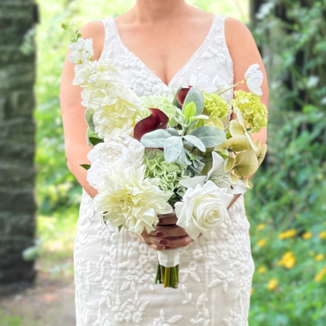 Bride holding a realistic artificial flower bridal bouquet featuring pale green artificial hydrangeas, white artificial dahlias, white artificial peonies, white artificial tea roses, white artificial Austin roses, and deep wine artificial calla lilies accented with soft lamb’s ear greenery — from My Faux Florist Canada.