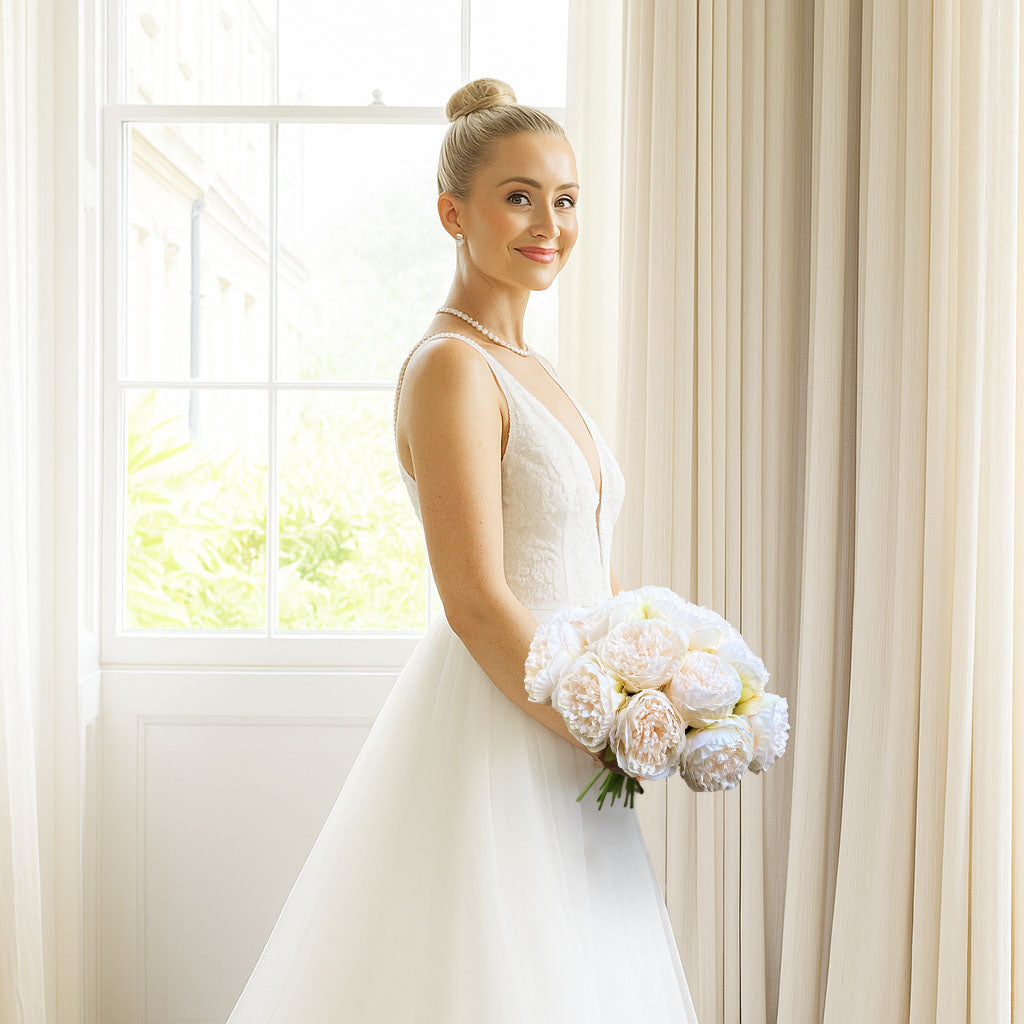 Bride holding a bouquet of white faux peonies in front of a cream curtain background, by My Faux Florist artificial flowers Canada.