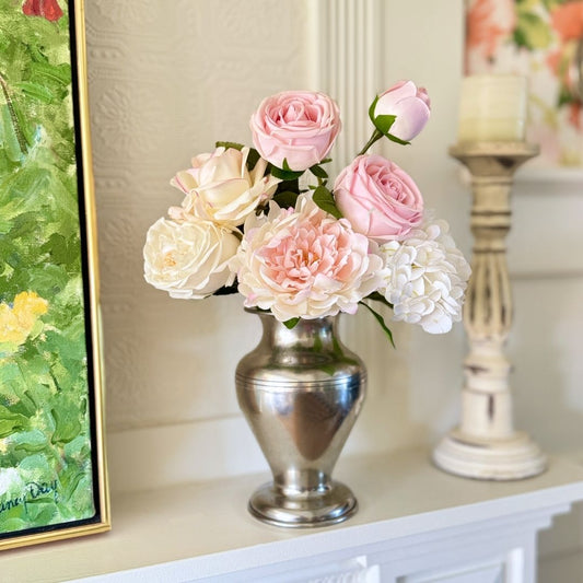 Silver vase containing realistic artificial flowers sits on a fireplace mantel.  The vase includes artificial roses, artificial peonies and artificial hydrangea in shades of pink, white and cream.  For My Faux Florist Canada.