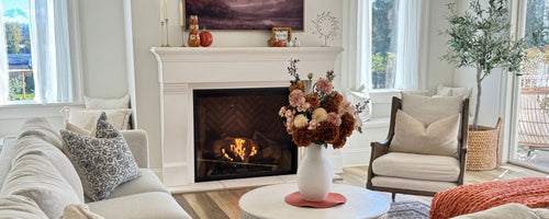 living room with fireplace in white and neutral colours. In the centre sits a large bouquet in a white vase of artificial flowers for Fall. Real Touch hydrangeas in deep orange, and artificial dahlias and Real Touch roses are visible. For My Faux Florist Canada.