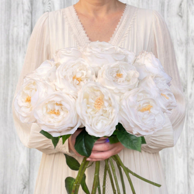 A person in a cream pleated dress holding a large bouquet of realistic white roses with green leaves and stems, Real Touch roses, posed against a light gray wood-plank background. For My Faux Florist Canada