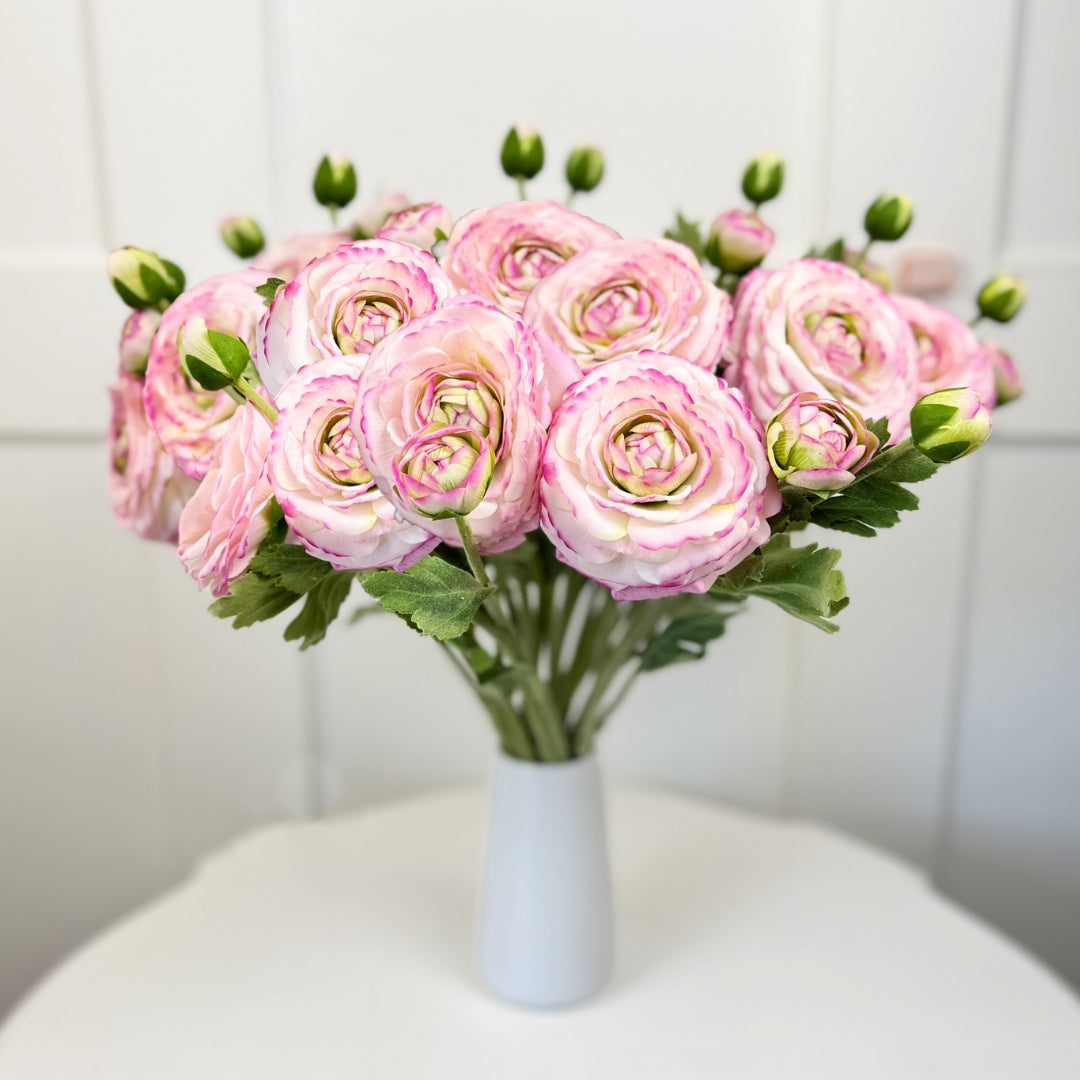 Bouquet of pink artificial ranunculus flowers in a white vase on a white surface with a white background