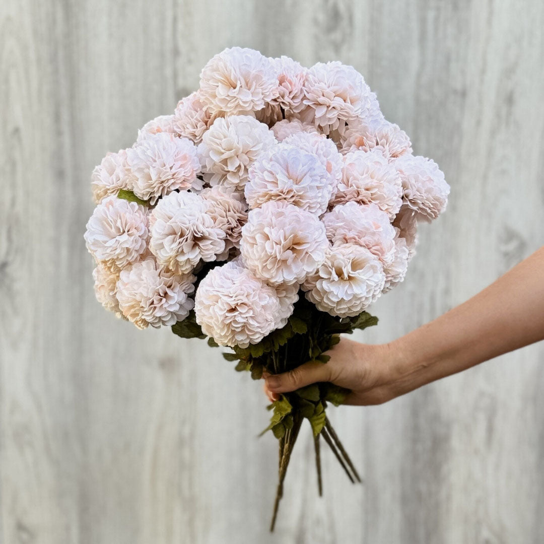 Person holding out a bunch of cream-colored artificial chrysanthemum flowers in front of a plain background, by My Faux Florist artificial flowers Canada.