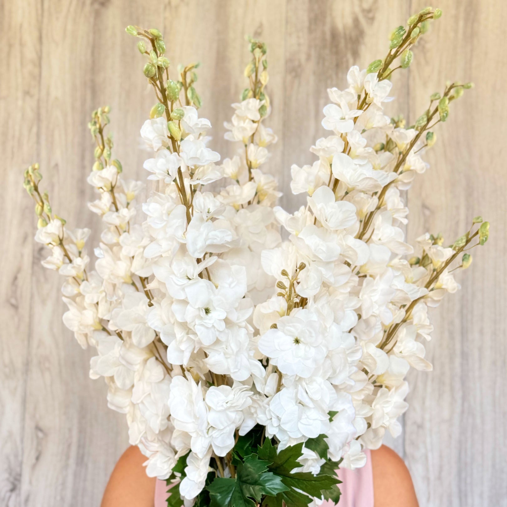 A bouquet of white artificial delphinium flowers with green leaves, held in front of a wooden background.