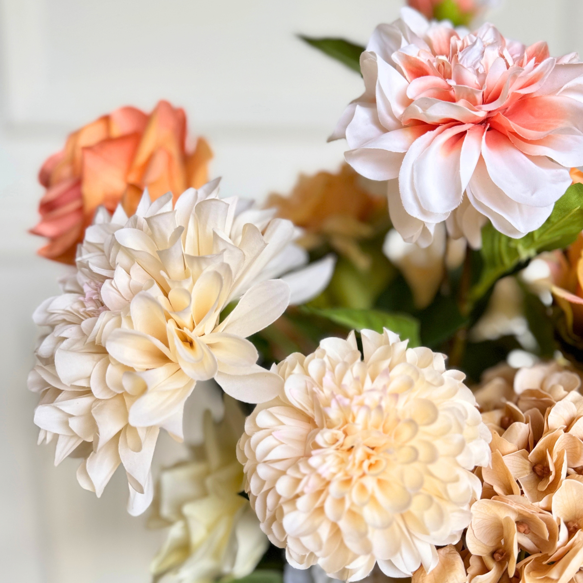 A close-up of realistic artificial flower stems arranged in a bouquet, featuring large cream dahlia blooms, a peach rose, and pale peach hydrangea-like flowers with green leaves against a soft light background. For My Faux Florist Canada.