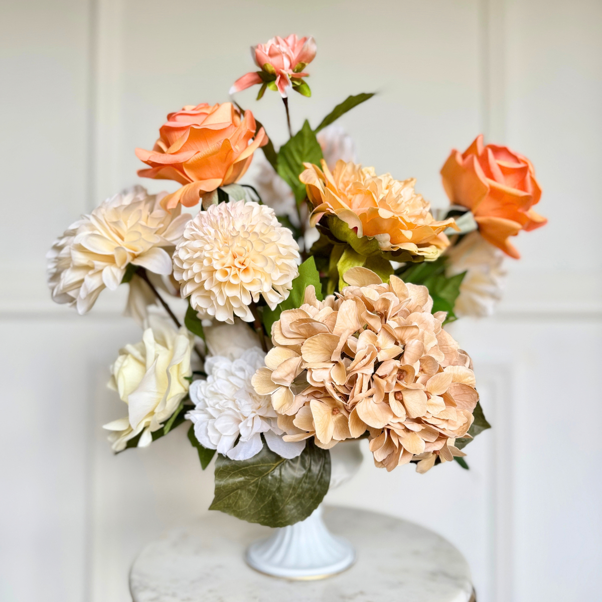 A realistic artificial flower arrangement featuring peach Real Touch roses, cream Real Touch dahlias, and beige Real Touch hydrangeas with green leaves, displayed in a white pedestal vase on a marble-topped surface against a white paneled wall. For My Faux Florist Canada.