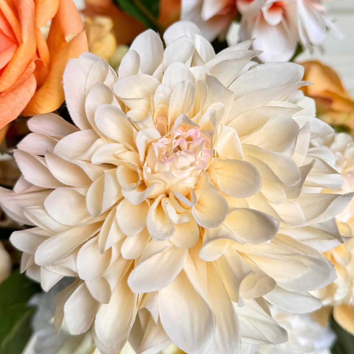 A close-up of a large cream realistic artificial dahlia. Real Touch dahlia with layered petals, surrounded by soft peach roses and hints of other flowers. For My Faux Florist Canada.