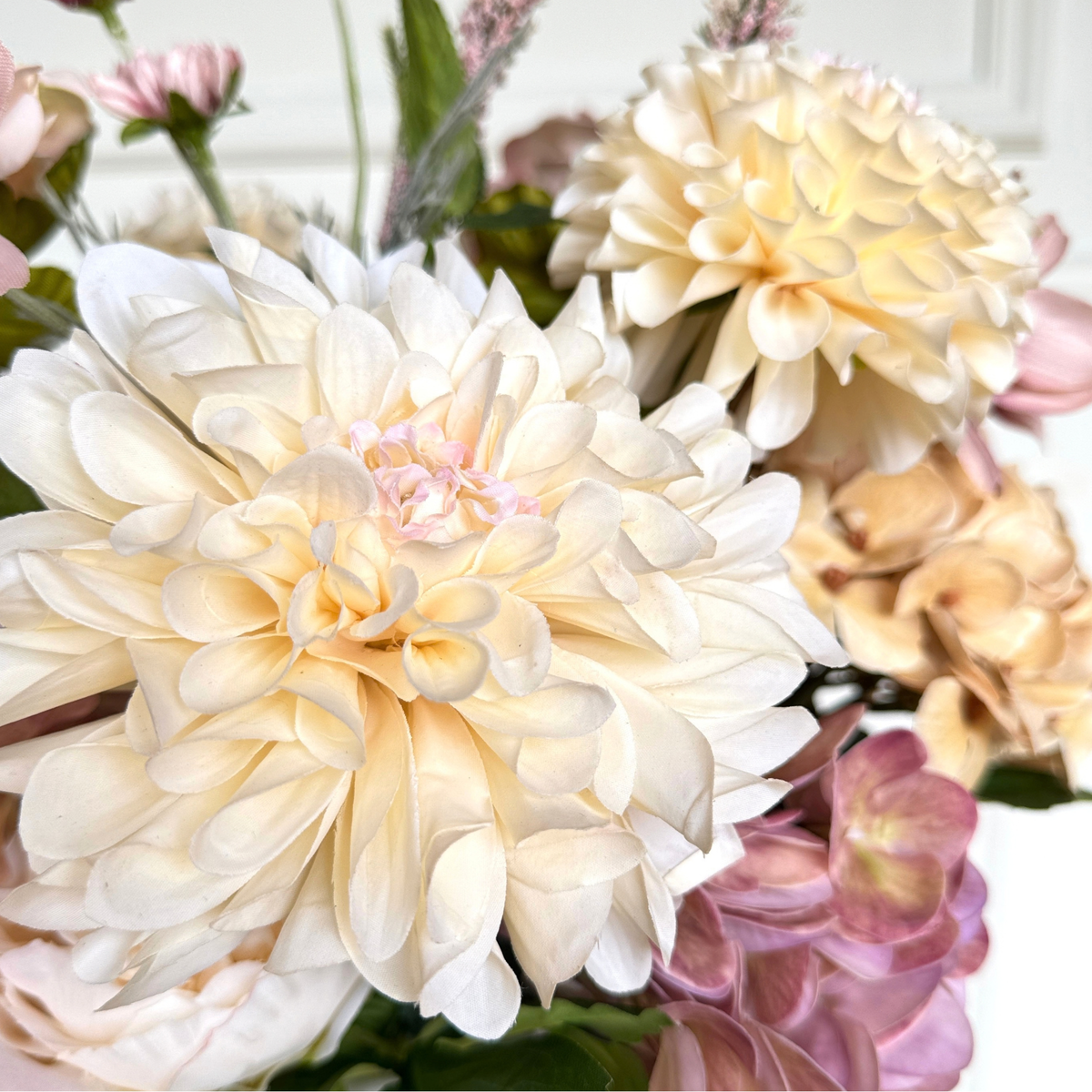 A close-up of a realistic faux flower arrangement featuring large cream and pale yellow artificial dahlia flowers with soft pink accents and some green foliage. For My Faux Florist Canada.