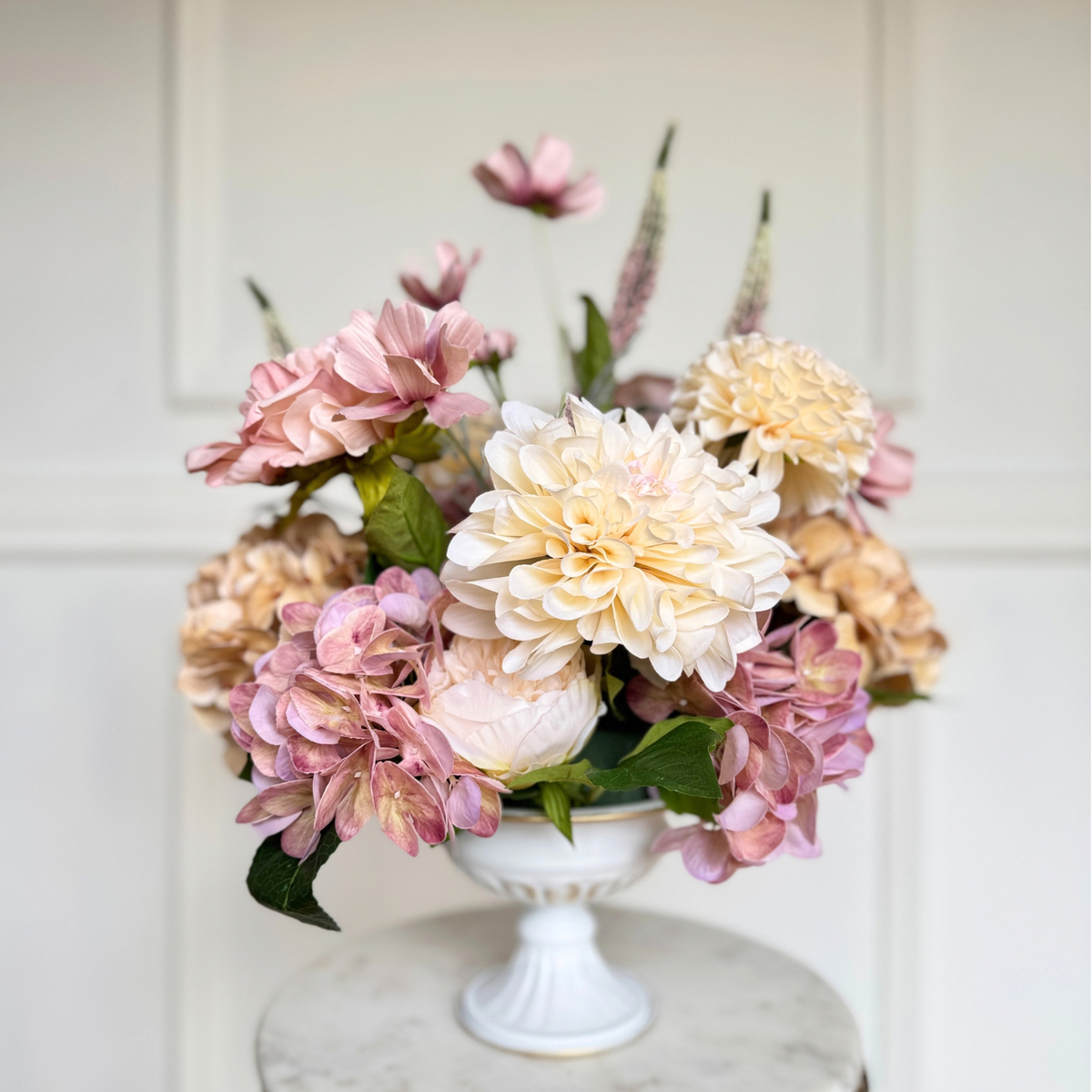 A realistic artificial flower arrangement of faux pink Real Touch hydrangeas, cream Real Touch peonies, and green foliage in a white pedestal bowl, set on a marble-topped table against a white paneled wall. For My Faux Florist Canada.