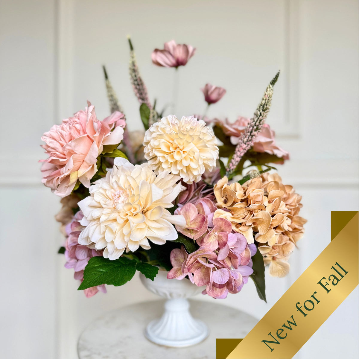 A faux floral arrangement in a white pedestal vase, featuring pink, cream, and peach flowers with green leaves, set against a light paneled wall. A gold diagonal banner in the lower right corner reads 'New for Fall'. For My Faux Florist Canada.