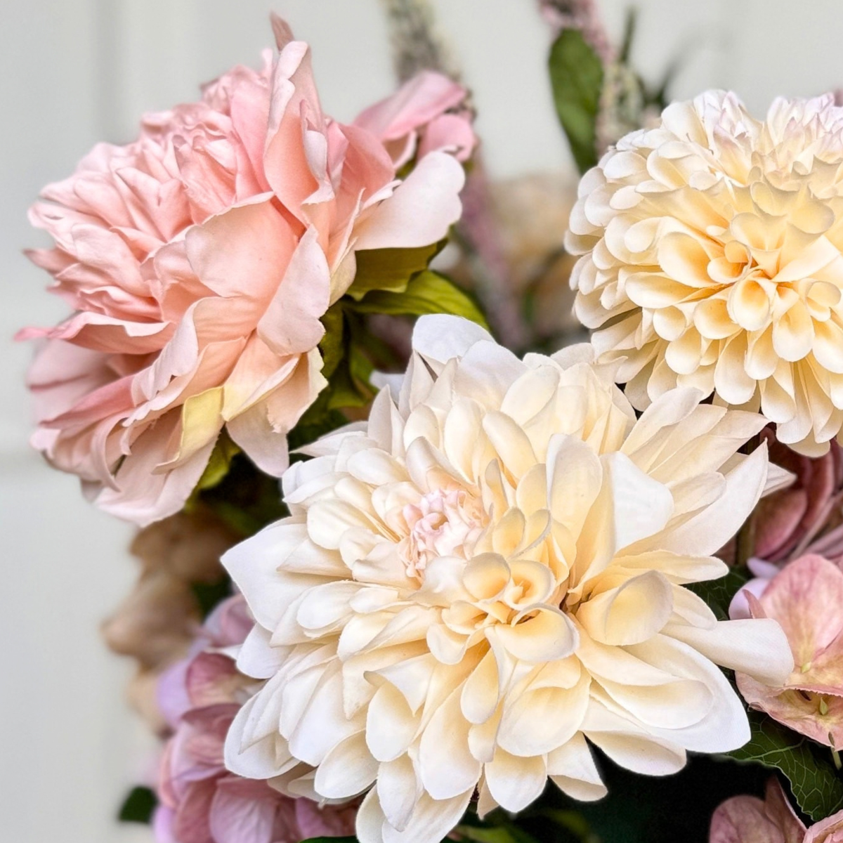 A close-up of realistic artificial flowers, featuring a large pale cream Real Touch dahlia in the foreground, a soft blush pink artificial peony on the left, and other muted pink blooms with green leaves against a light neutral background. For My Faux Florist Canada.