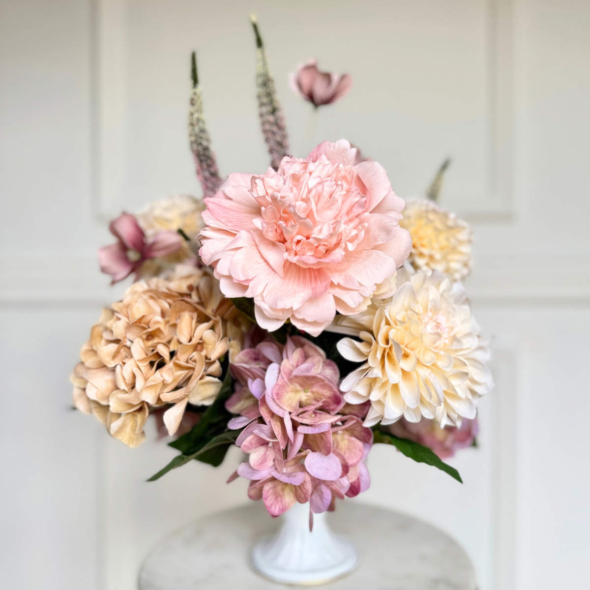A bouquet of realistic artificial flowers in soft pastel tones, including a large pink faux peony, cream and blush Real Touch hydrangea-like blooms, and muted green stems, arranged in a small white pedestal vase against a light paneled wall. For My Faux Florist Canada.
