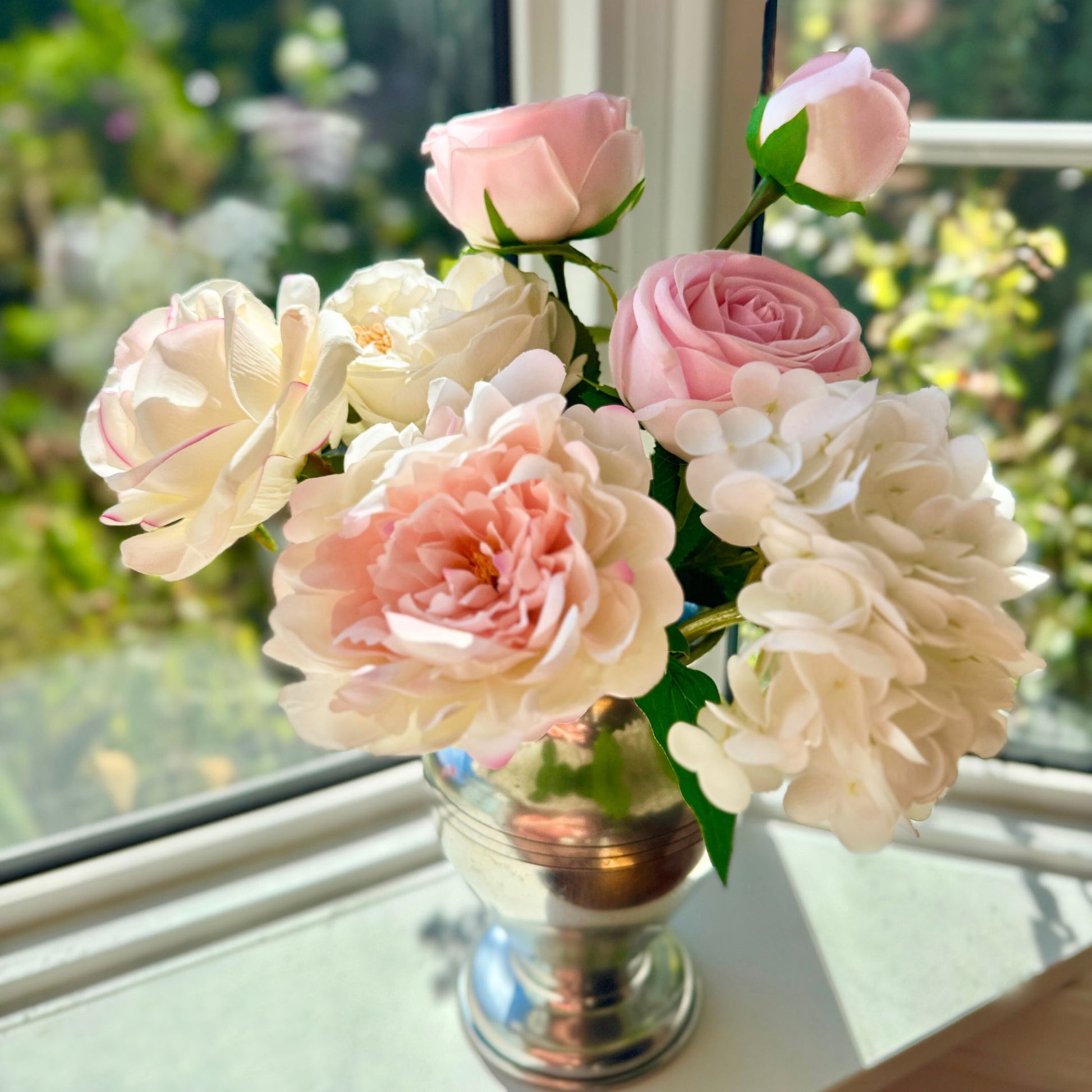 Silver vase containing realistic artificial flowers sits on a windowsill.  The vase includes artificial roses, artificial peonies and artificial hydrangea in shades of pink, white and cream.  For My Faux Florist Canada.