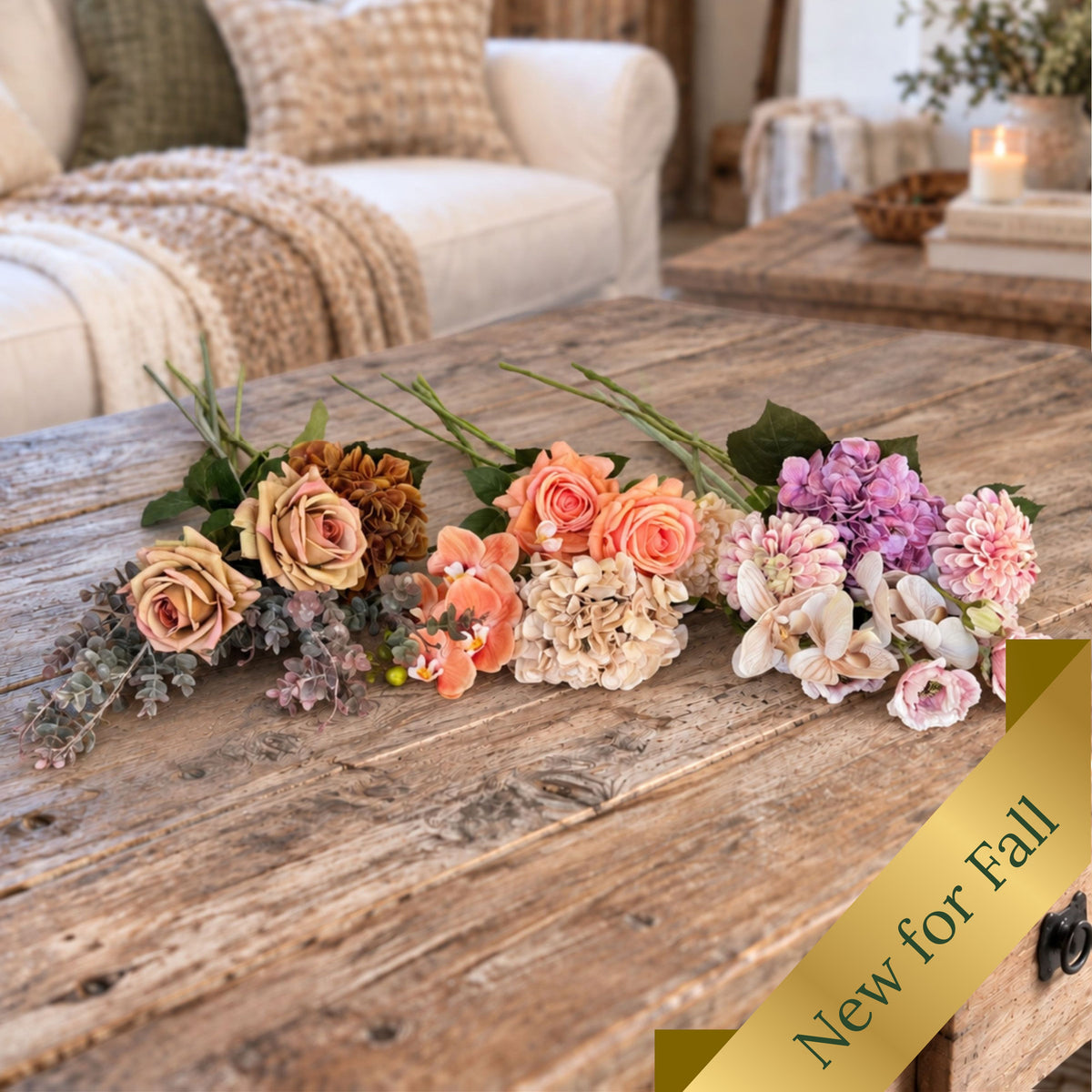 A rustic wooden table in a cozy living room setting displays a group of three bunches of realistic artificial flowers.