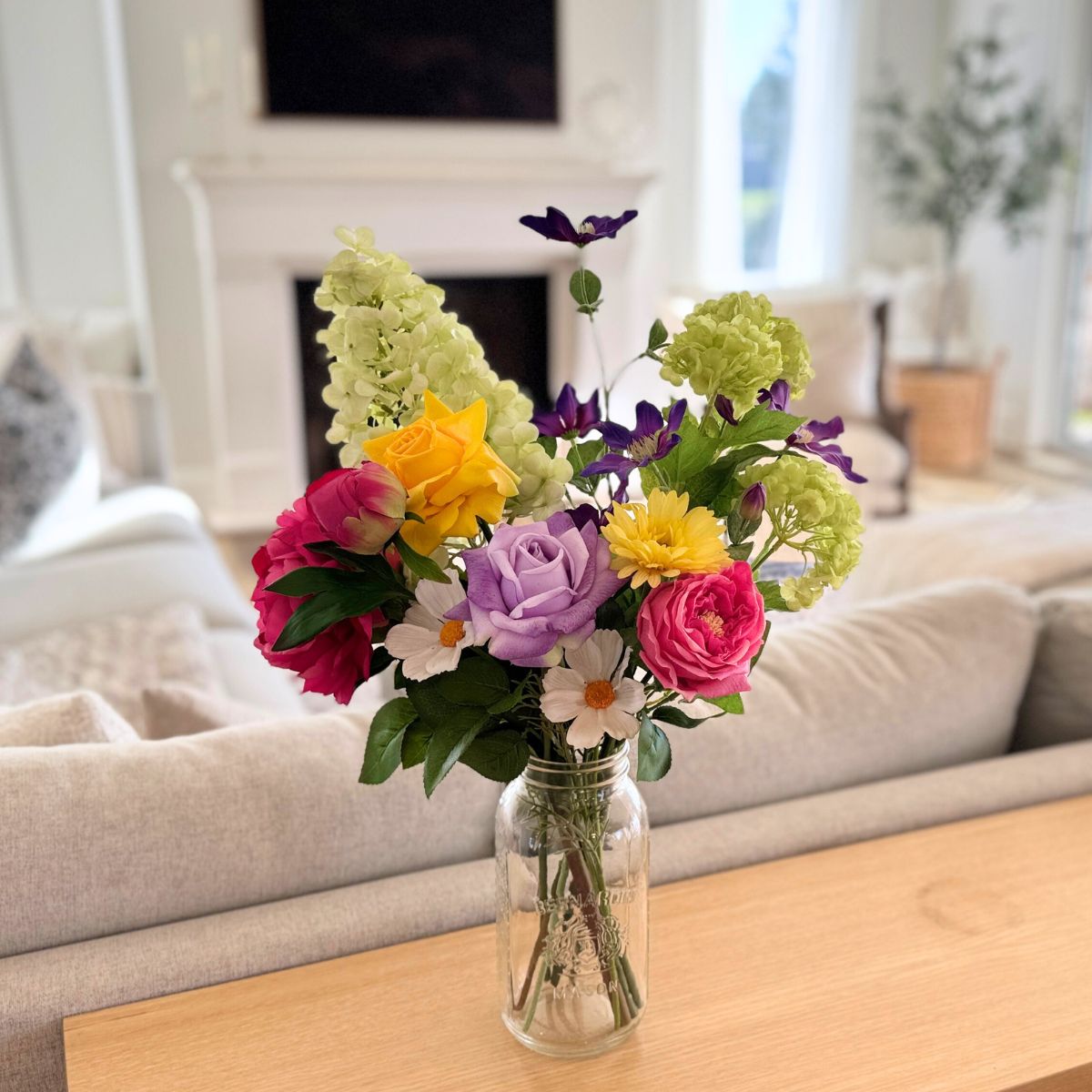 Colorful bouquet of realistic artificial flowers in a glass vase on a wooden table in a living room. For My Faux Florist Canada.