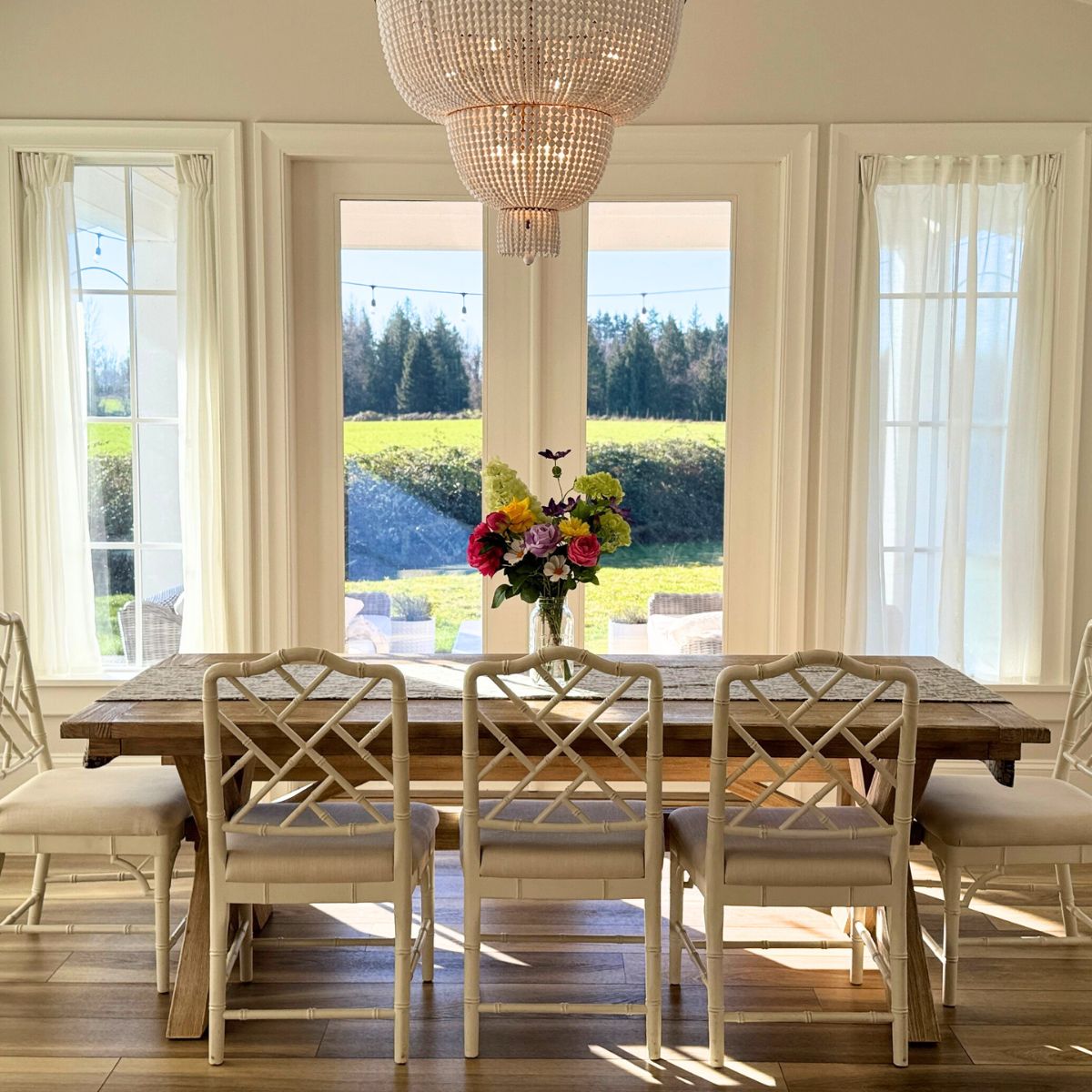 Dining room with wooden table and chairs, large windows, and a chandelier. On the table is a realistic artificial flower arrangement in bright colours,  for My Faux Florist Canada.