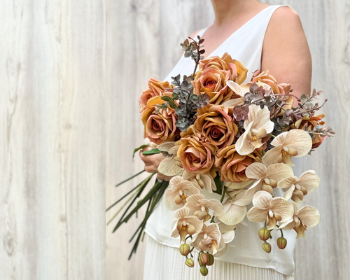 A person in a white sleeveless dress holding a large bouquet of realistic artificial flowers for fall in orange, tan, cream, and muted brown flowers against a light wood plank wall. for My Faux Florist Canada.