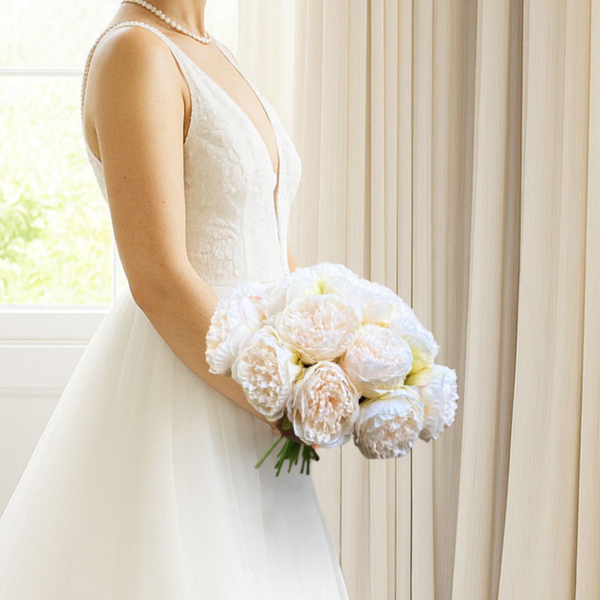 Bride holding a bouquet of white faux peonies in front of a cream curtain, by My Faux Florist artificial flowers Canada.