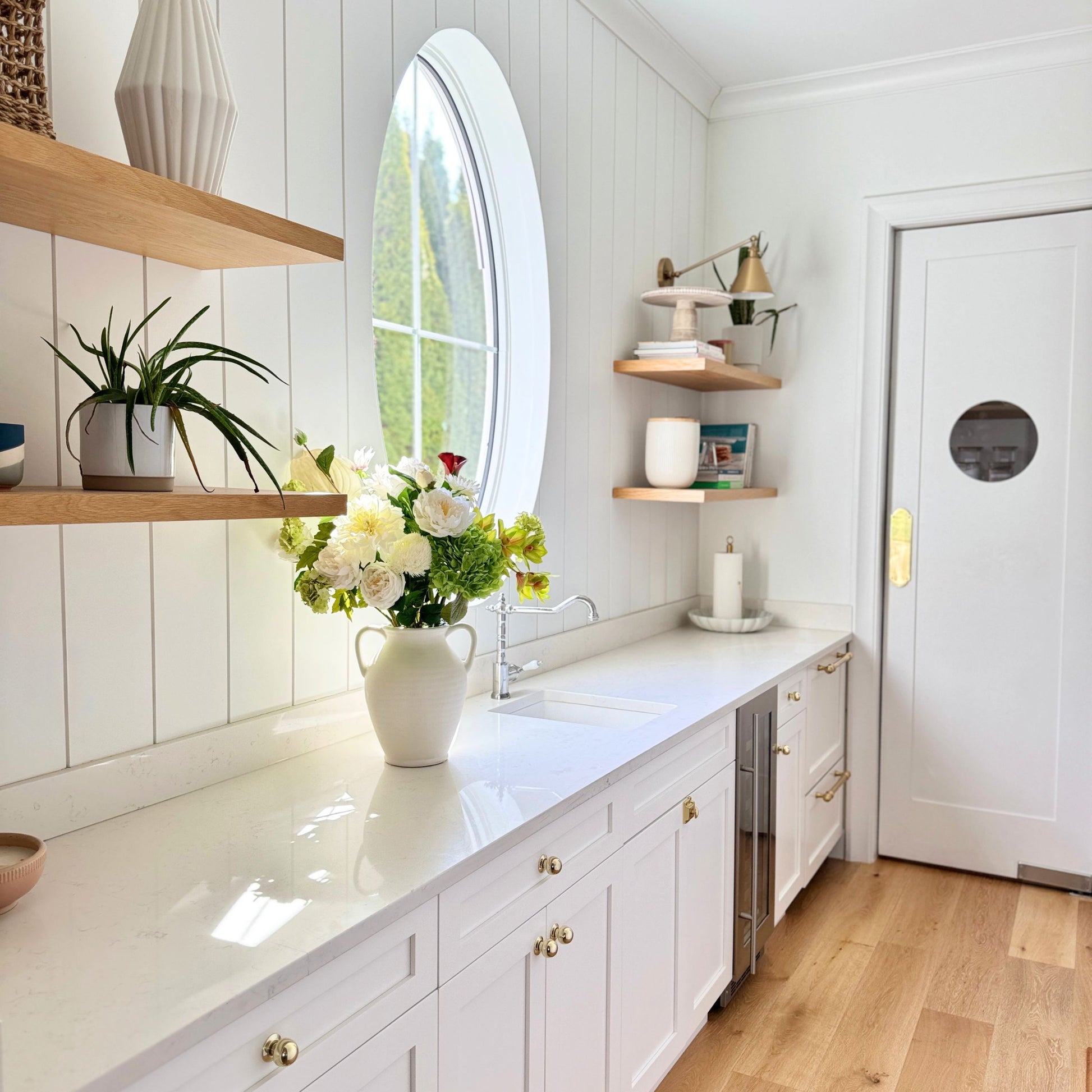 Modern kitchen with white countertops, cabinets, a white vase of realistic artificial flowers, and a round mirror. Faux peonies, faux roses, faux hydrangea, and faux orchids in an artificial flower bouquet for My Faux Florist Canada.