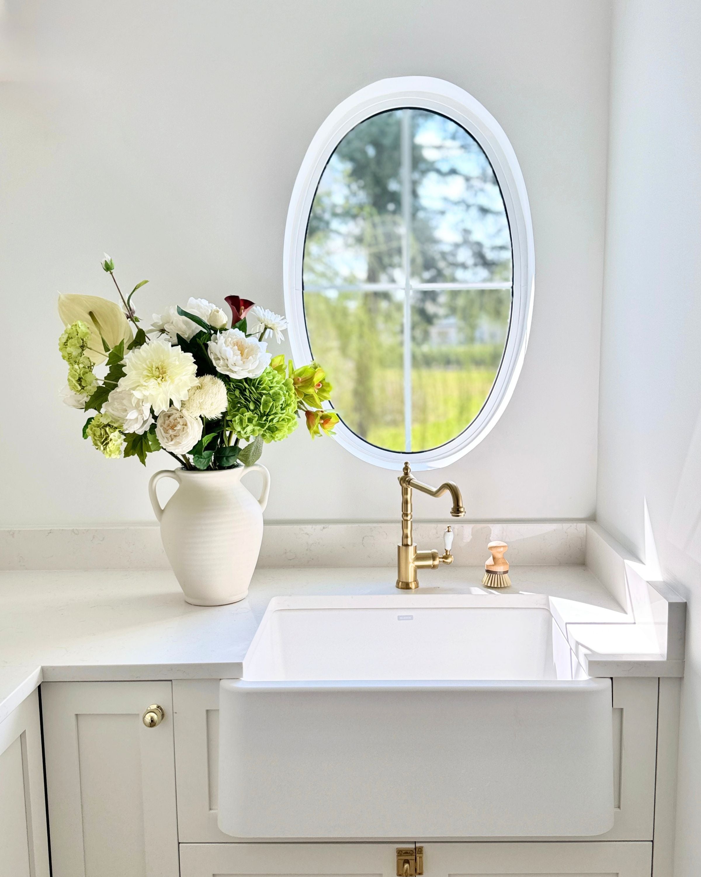 A vase of white and green realistic artificial flowers sits on a counter next to a small whit sink and oval window. Faux peonies, faux roses, faux hydrangea, and faux orchids in an artificial flower bouquet for My Faux Florist Canada.