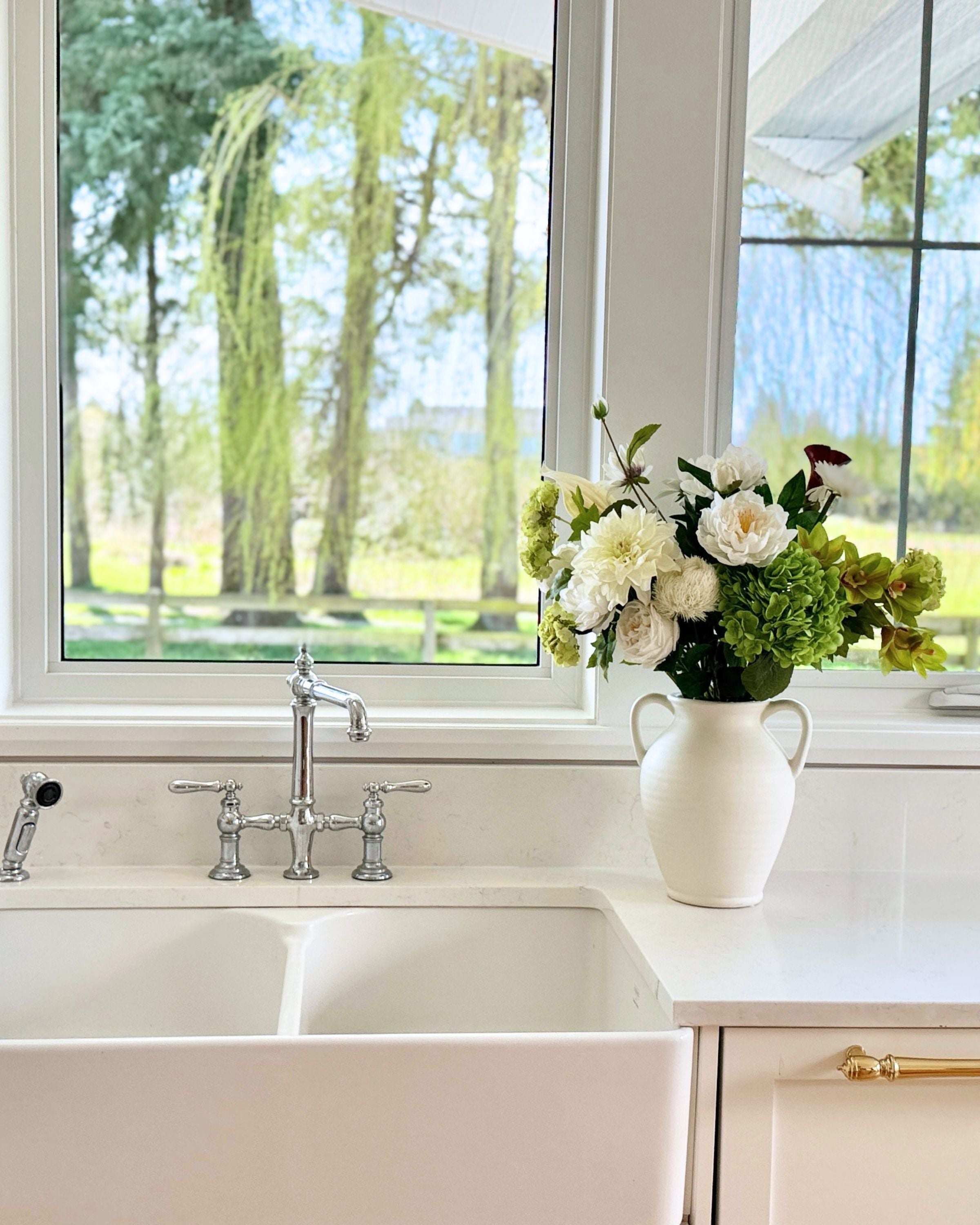 Kitchen sink with a vase of realistic artificial  flowers on the counter, large window in the background. Faux peonies, faux roses, faux hydrangea, and faux orchids in an artificial flower bouquet for My Faux Florist Canada.