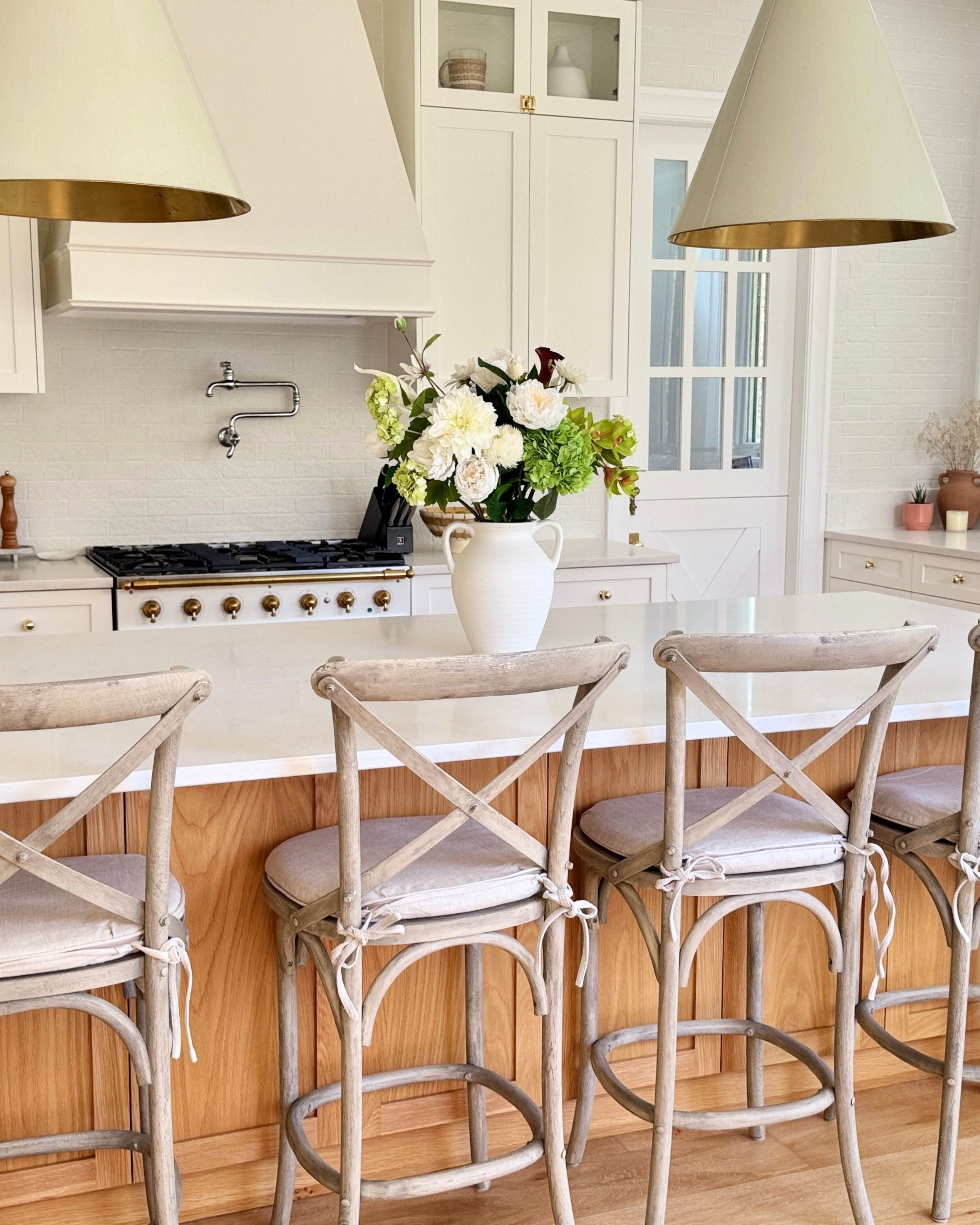 Kitchen with white cabinets, wooden floor, and bar stools with white cushions.  A large vase of white and green realistic artificial flowers is on the counter. Faux peonies, faux roses, faux hydrangea, and faux orchids in an artificial flower bouquet for My Faux Florist Canada.